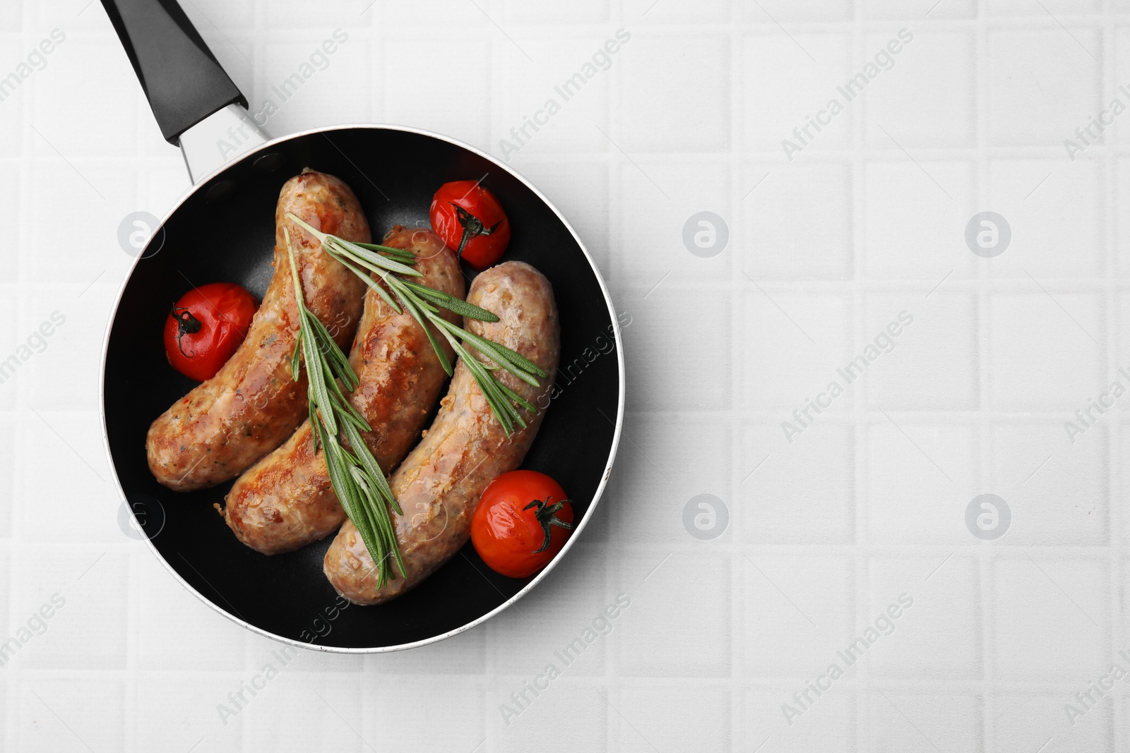 Photo of Frying pan with tasty homemade sausages, rosemary and tomatoes on white tiled table, top view. Space for text