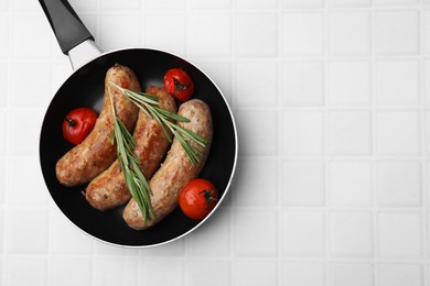 Frying pan with tasty homemade sausages, rosemary and tomatoes on white tiled table, top view. Space for text