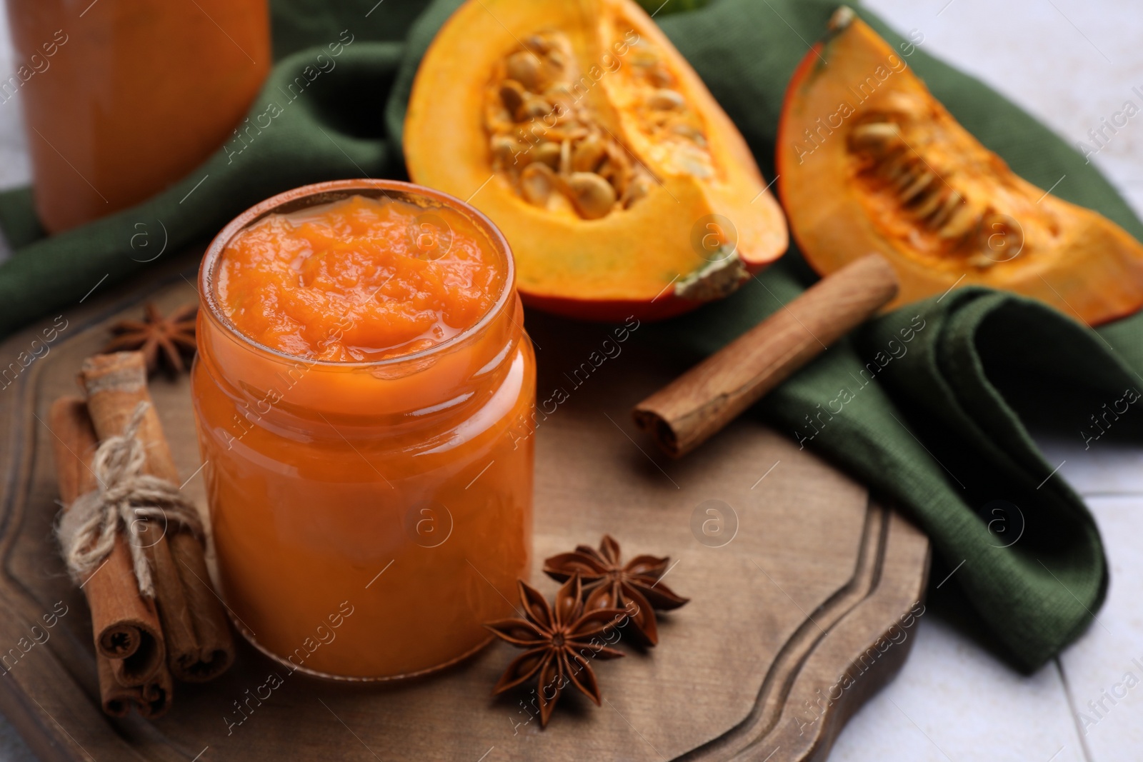 Photo of Jar of delicious pumpkin jam and ingredients on tiled surface, closeup