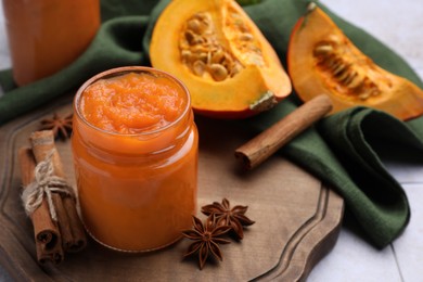 Photo of Jar of delicious pumpkin jam and ingredients on tiled surface, closeup