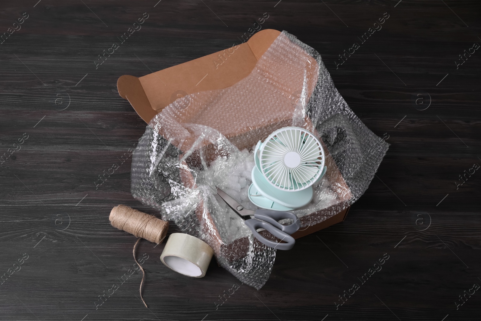 Photo of Small fan with bubble wrap in cardboard box and packaging items on dark wooden table, above view