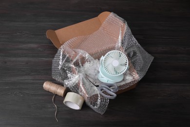 Small fan with bubble wrap in cardboard box and packaging items on dark wooden table, above view