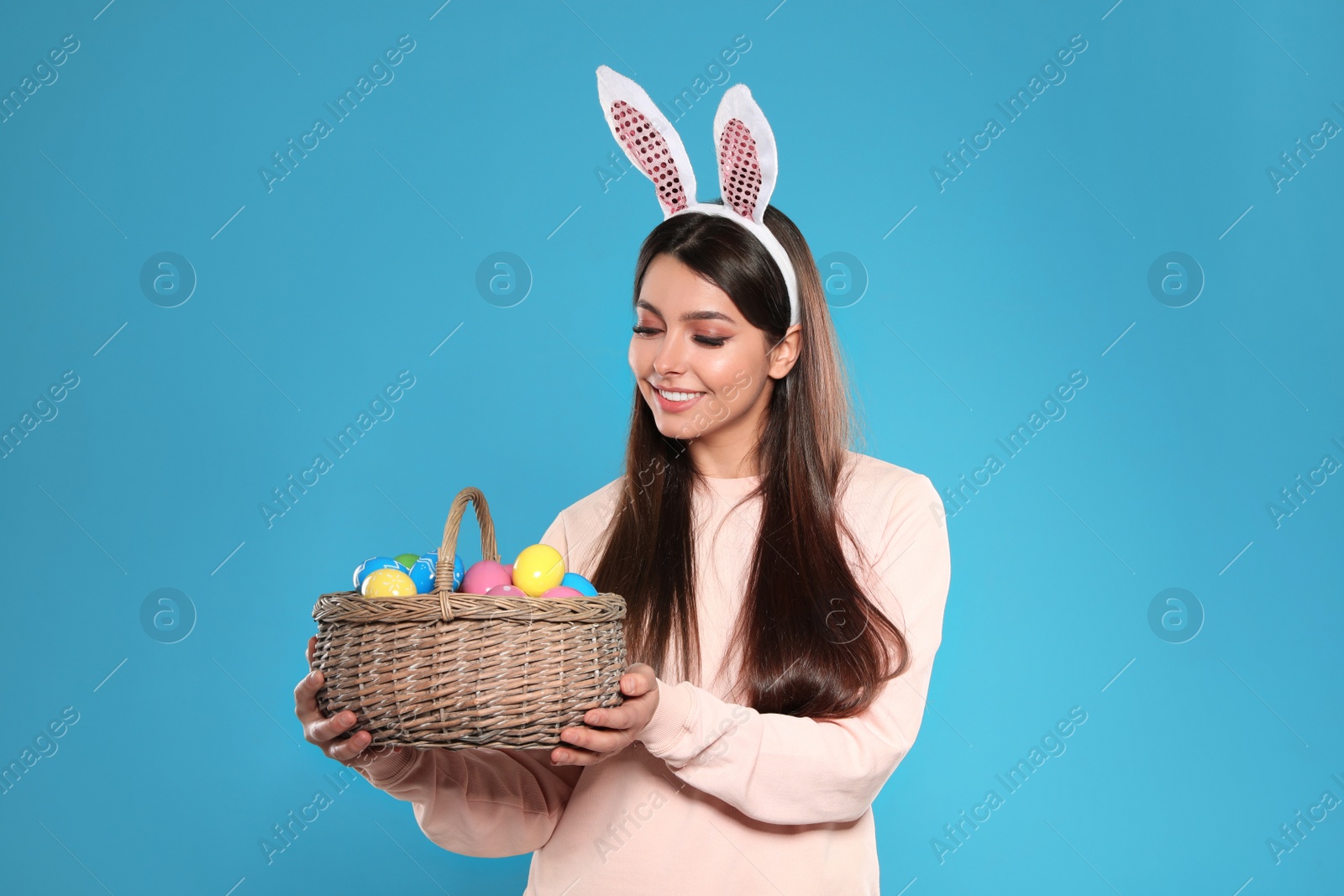 Photo of Beautiful woman in bunny ears headband holding basket with Easter eggs on color background