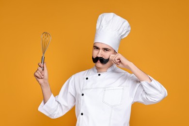 Portrait of happy confectioner with funny artificial moustache holding whisk on orange background