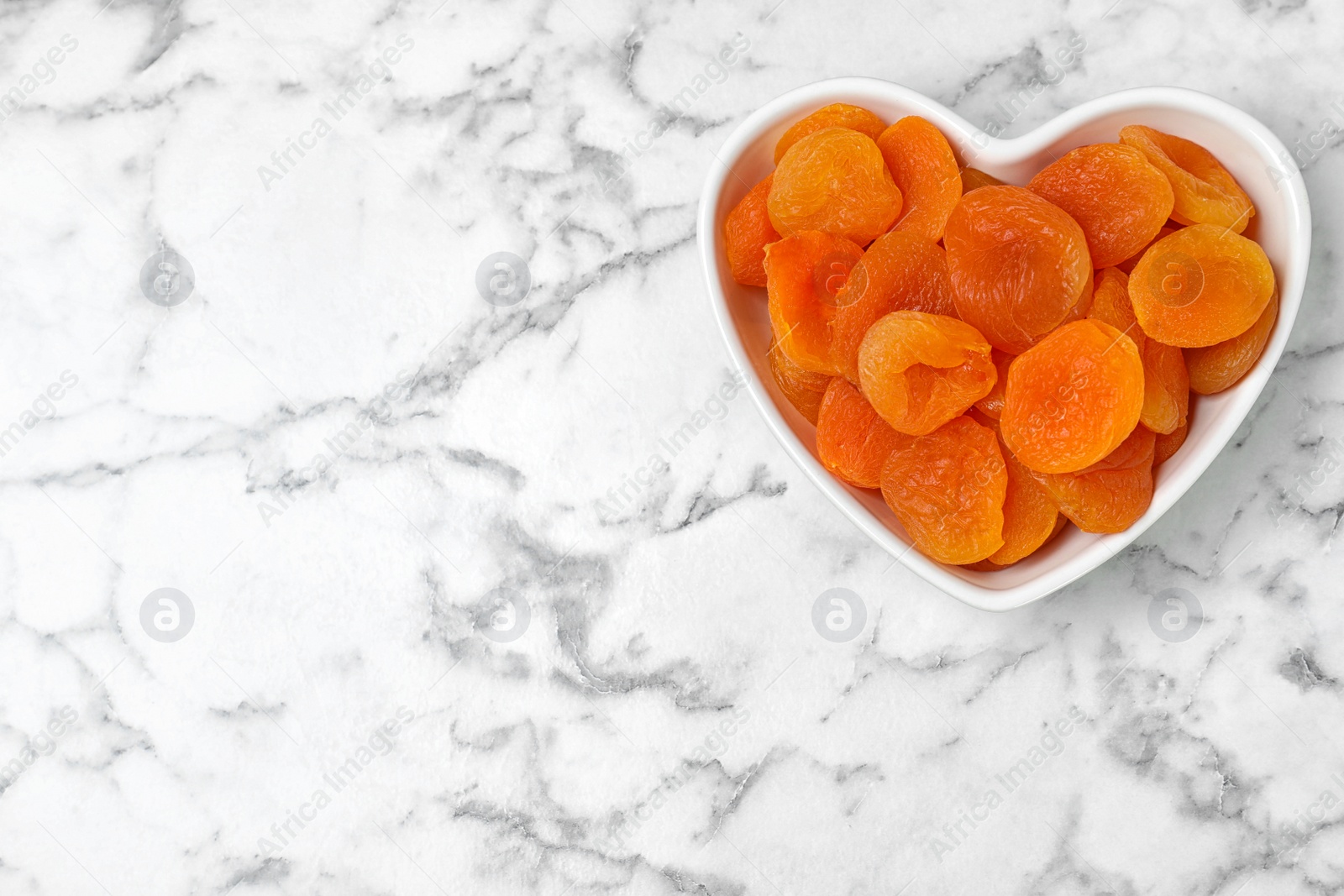 Photo of Heart shaped bowl of apricots on marble background, top view with space for text. Dried fruit as healthy food