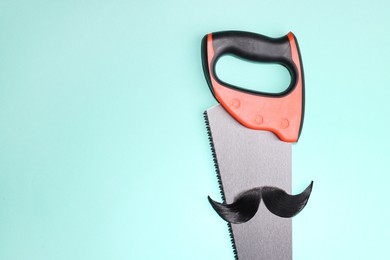 Man's face made of artificial mustache and hand saw on light blue background, top view. Space for text