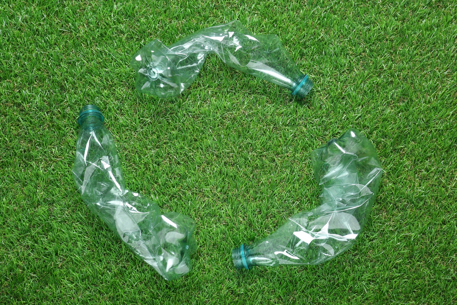 Photo of Recycling symbol made of crumpled plastic bottles on green grass, flat lay
