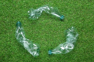 Photo of Recycling symbol madecrumpled plastic bottles on green grass, flat lay