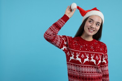 Happy young woman in Christmas sweater and Santa hat on light blue background. Space for text