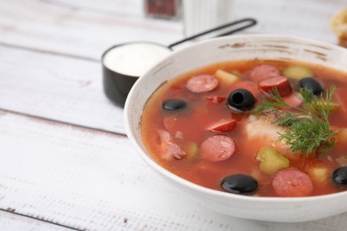 Photo of Meat solyanka soup with thin dry smoked sausages in bowl on white wooden table, closeup. Space for text