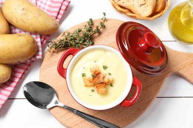 Photo of Tasty potato soup with croutons and rosemary in ceramic pot served on white wooden table, flat lay