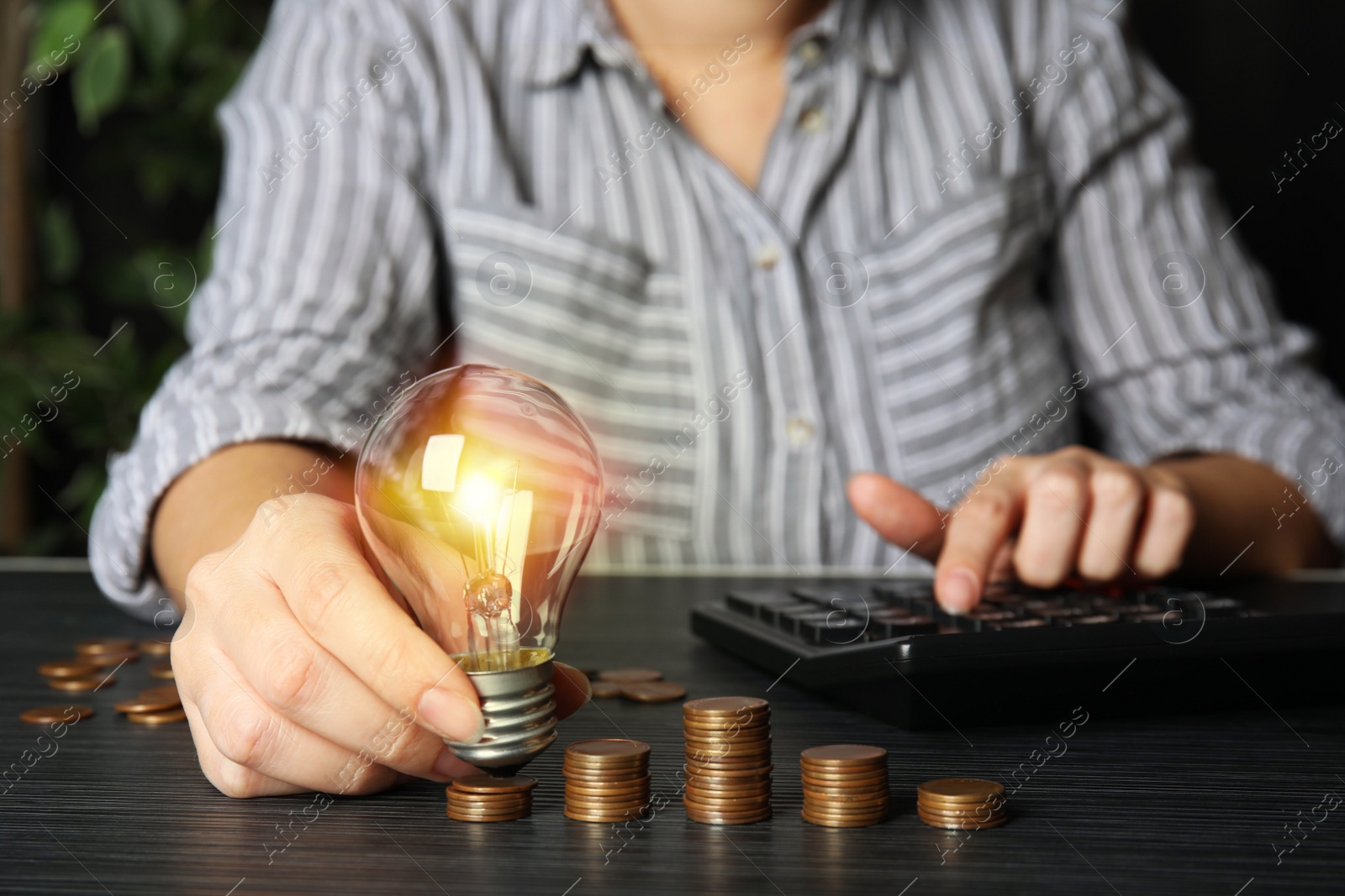 Photo of Woman with light bulb, calculator and coins at black wooden table, closeup. Power saving