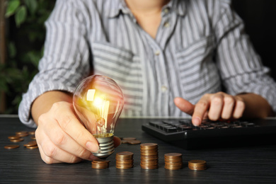 Photo of Woman with light bulb, calculator and coins at black wooden table, closeup. Power saving
