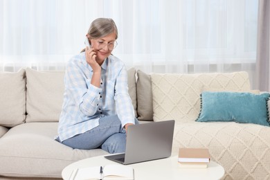 Photo of Beautiful senior woman talking on phone while using laptop at home