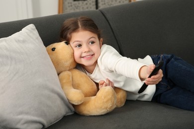 Photo of Little girl with toy bear changing TV channels by remote control on sofa at home