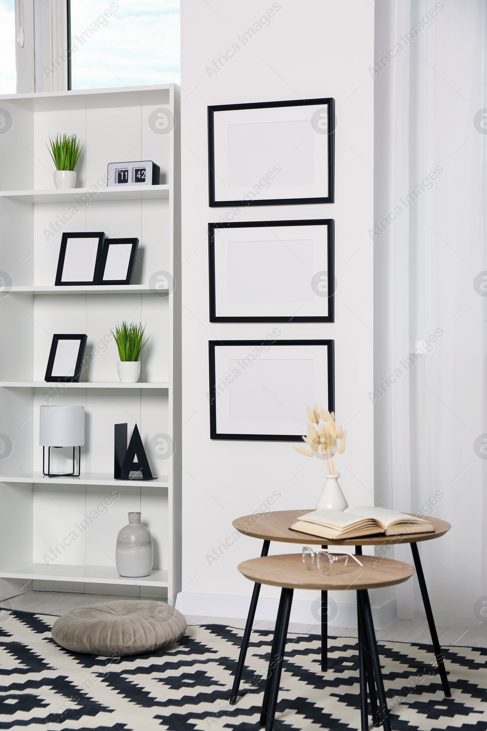 Photo of Stylish room interior with empty frames hanging on white wall near shelving unit