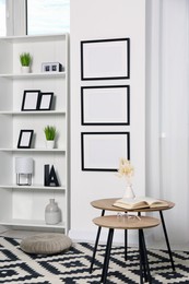 Photo of Stylish room interior with empty frames hanging on white wall near shelving unit