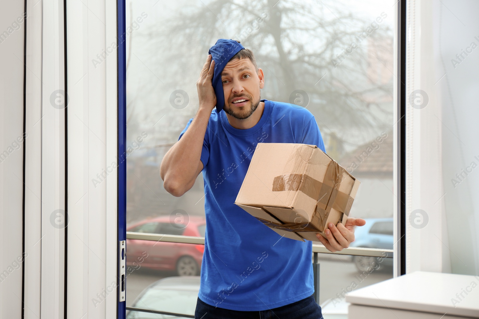 Photo of Emotional courier with damaged cardboard box in doorway. Poor quality delivery service