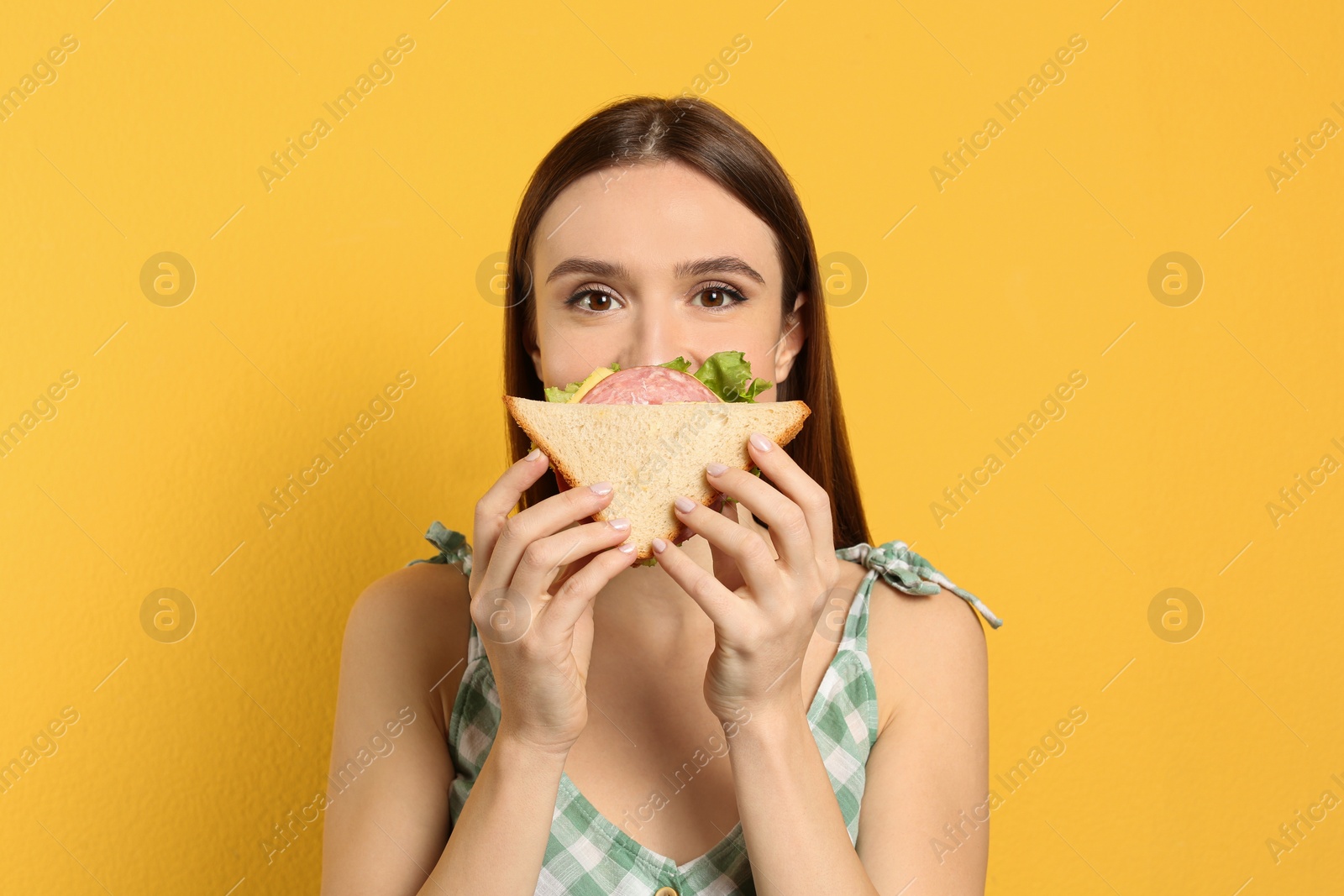 Photo of Young woman eating tasty sandwich on yellow background