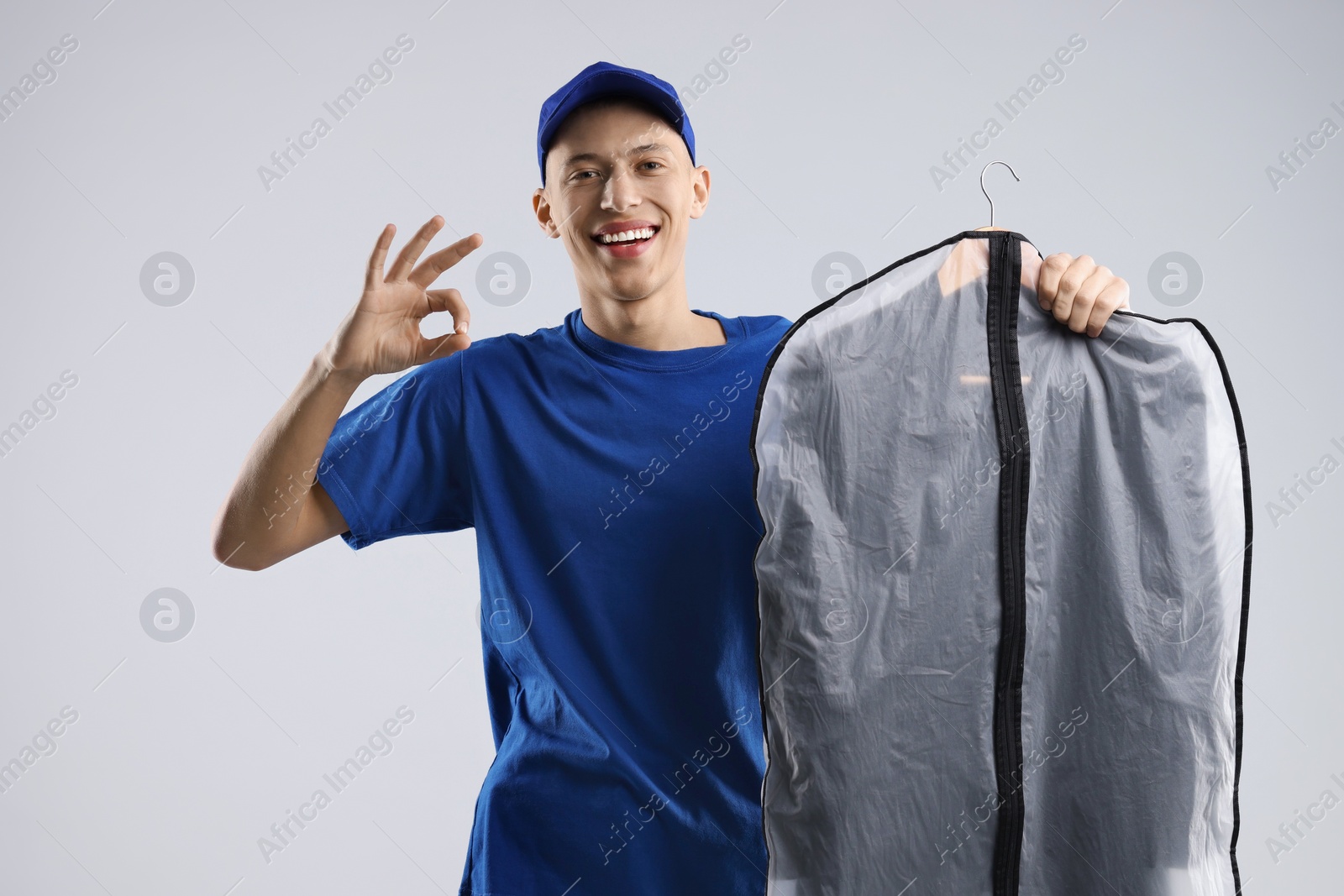 Photo of Dry-cleaning delivery. Happy courier holding garment cover with clothes and showing OK gesture on light grey background