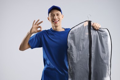 Dry-cleaning delivery. Happy courier holding garment cover with clothes and showing OK gesture on light grey background