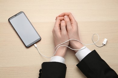 Photo of Man showing hands tied to smartphone with charging cable and at wooden table, top view. Internet addiction
