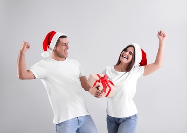 Beautiful happy couple in Santa hats holding Christmas gift on light background