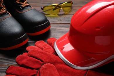 Photo of Hard hat, pair of working boots, protective gloves and goggles on wooden background, closeup