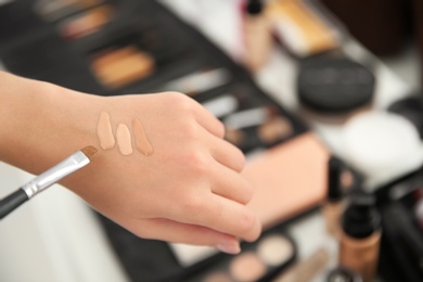 Photo of Woman testing different shades of liquid foundation on her hand against blurred background, closeup