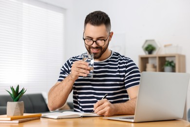 Young man with glass of water writing in notebook at wooden table indoors
