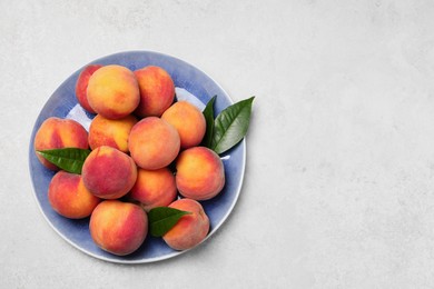 Many whole fresh ripe peaches and green leaves in plate on white table, top view. Space for text
