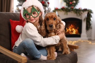 Photo of Cute little girl in Santa hat and Christmas glasses with English Cocker Spaniel at home