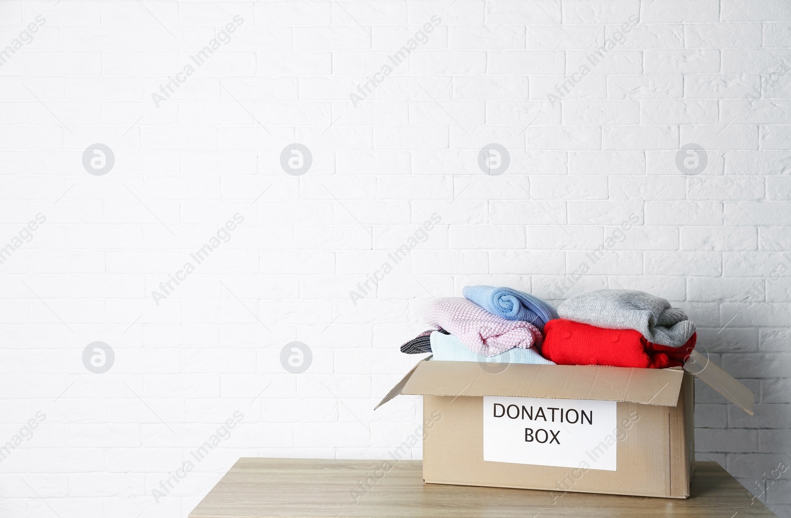 Photo of Donation box with knitted clothes on table near brick wall. Space for text