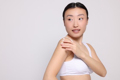Beautiful young Asian woman applying body cream onto shoulder on light grey background, space for text