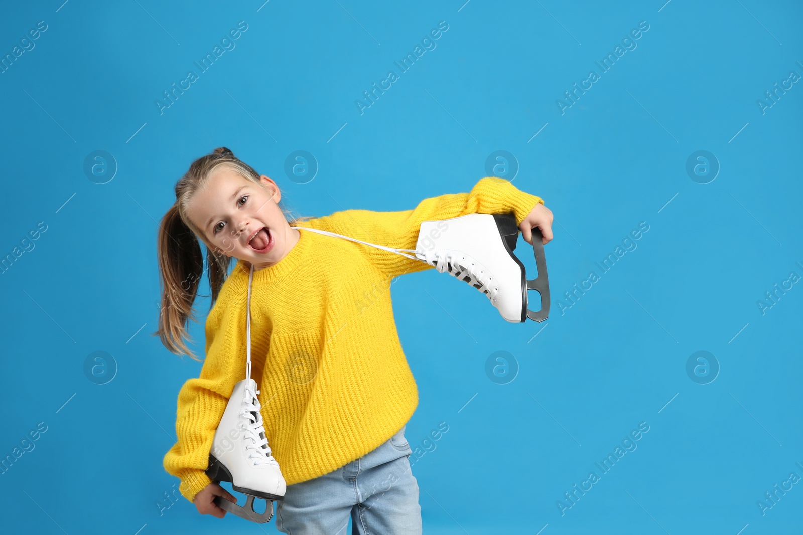 Photo of Excited little girl in yellow knitted sweater with ice skates on blue background