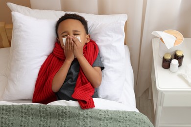 Photo of African-American boy with scarf and tissue blowing nose in bed indoors, above view. Cold symptoms