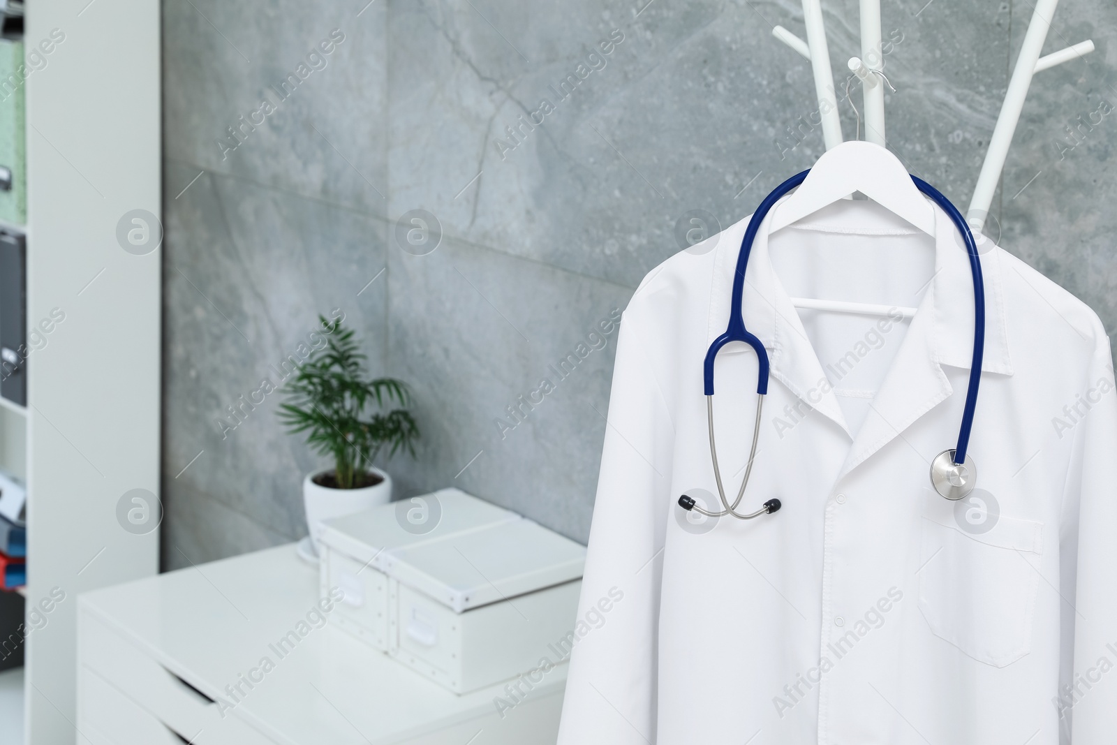 Photo of White doctor's gown and stethoscope hanging on rack in clinic, closeup. Space for text