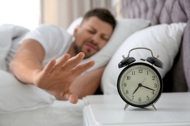 Photo of Sleepy man turning off alarm clock at home in morning, focus on hand
