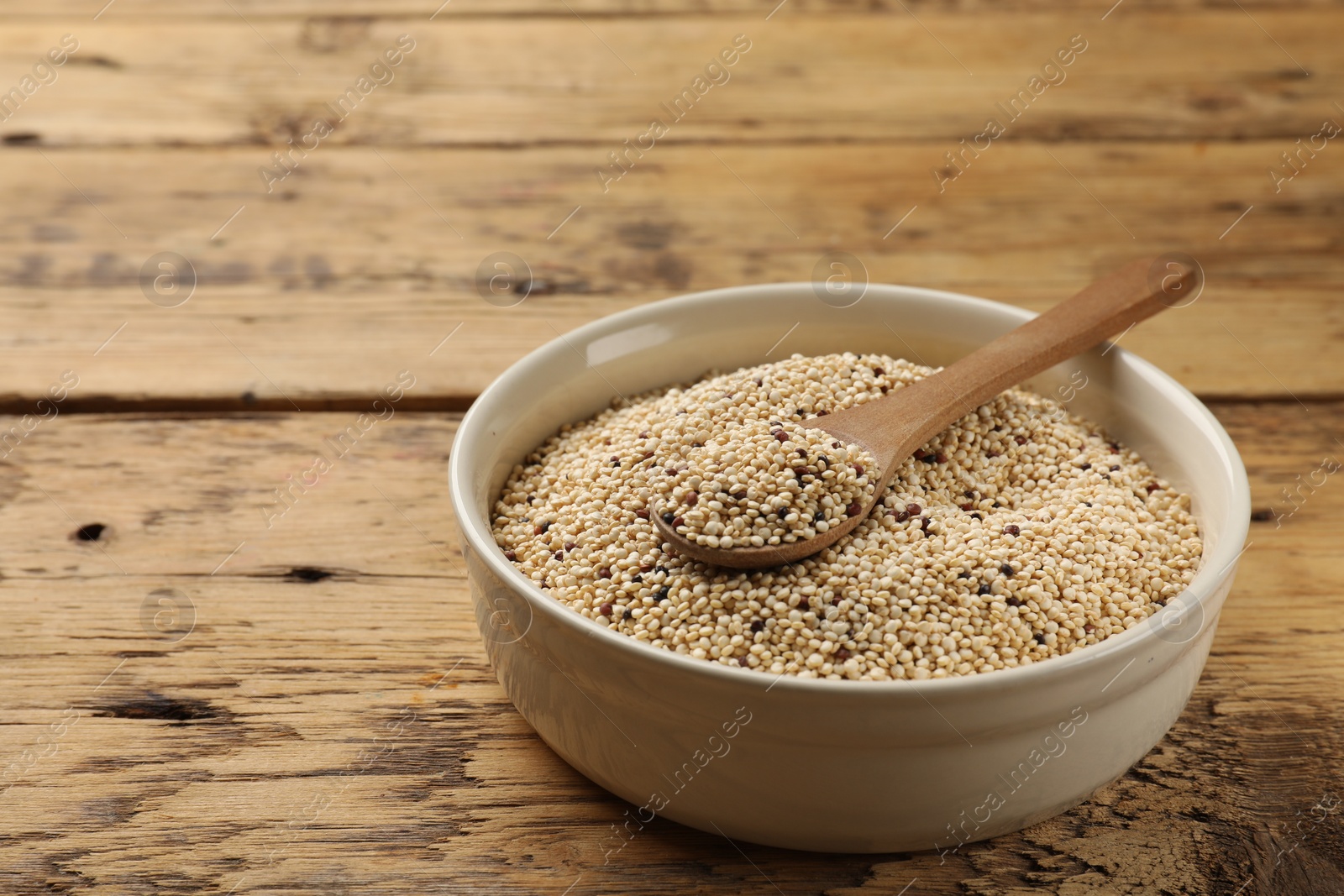 Photo of Raw quinoa seeds and spoon in bowl on wooden table, closeup. Space for text