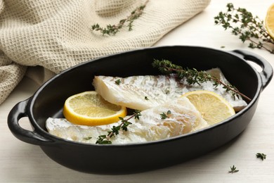 Photo of Fresh raw cod fillets with thyme and lemon in baking dish on white wooden table