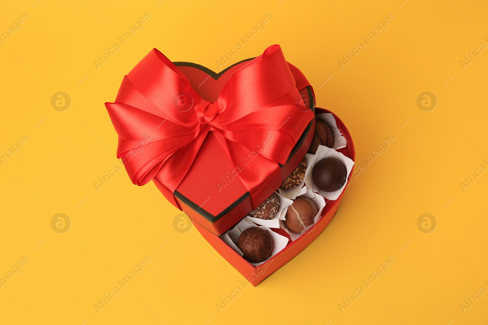 Photo of Heart shaped box with delicious chocolate candies on yellow background, above view