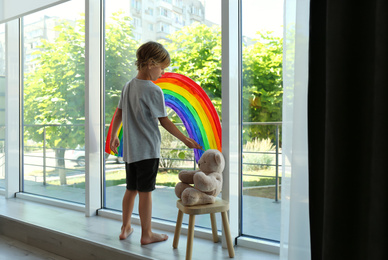 Photo of Little boy drawing rainbow on window indoors. Stay at home concept