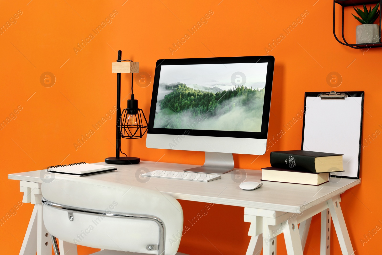 Photo of Modern computer, books, lamp and notebook on wooden desk near orange wall. Home office