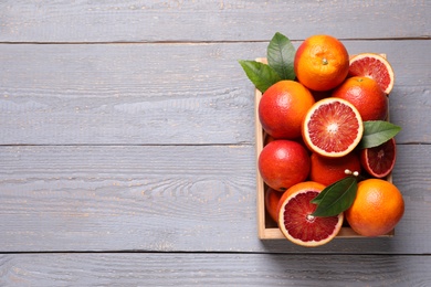 Photo of Crate of ripe red oranges and green leaves on grey wooden table, top view. Space for text