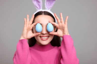 Happy woman in bunny ears headband holding painted Easter eggs near her eyes on grey background