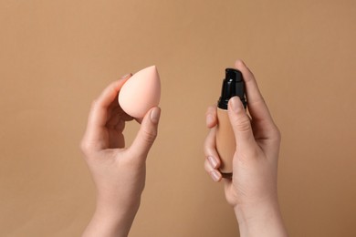 Photo of Woman holding bottle with skin foundation and makeup sponge on light brown background, closeup