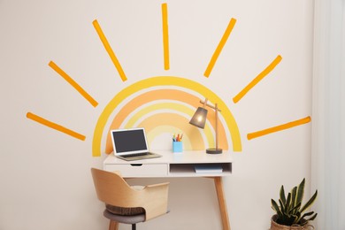 Laptop, stationery and lamp on white table near wall with painted sun indoors. Interior design