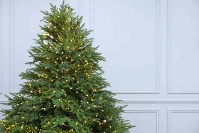 Beautiful Christmas tree decorated with festive lights near white wall indoors, space for text