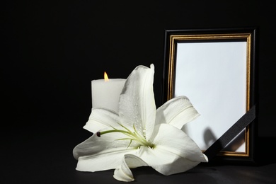 Funeral photo frame with ribbon, white lily and candle on dark table against black background. Space for design