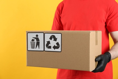 Photo of Courier holding cardboard box with different packaging symbols on yellow background, closeup. Parcel delivery